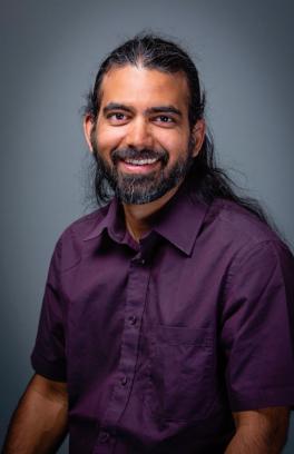Sachin Nedungadi, a brown-skinned man with long black hair and a full beard, is smiling in front of a grey-blue background and wearing a purple button-down.