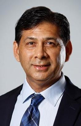 Irfan Rahman, a tan skinned man with short black hair, is wearing a suit and tie in a professional headshot with a grey background.