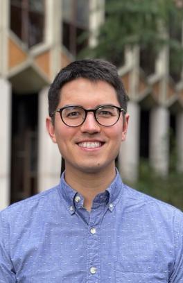 Douglas Reed, a light skinned man with short black hair and glasses, is standing in front of a blurred background of a triangular patterned building and smiling.