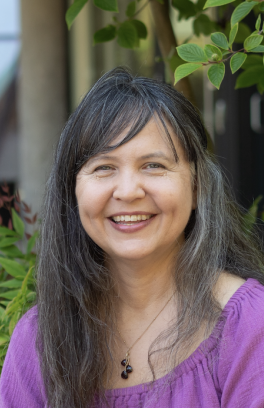 Ashlie Sarsgard, a light skinned older woman with black and grey hair and wearing a purple shirt, smiles in front of Portland State's greenery.