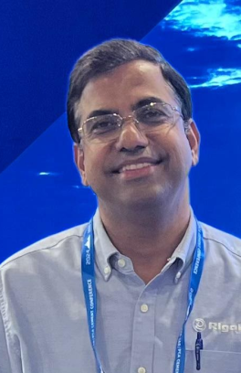 Akhilesh Tripathi, a brown-skinned man with short black hair and glasses, is smiling in front of a blue watery background.