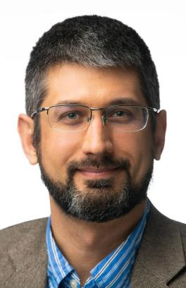 Eric Wiedner, a light-skinned man with a shirt dark hair and a beard, smiling in front of a white background. 