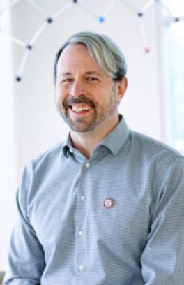 Matthew Sigman, a white man with short grey and black hair, smiling in front of a blurred background with chemical structures.