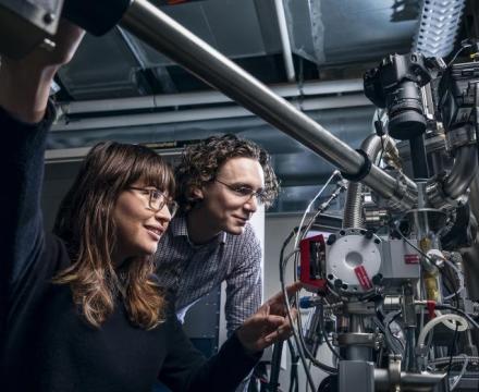 Two students adjusting and viewing a metal object, presumably a semiconductor, which consists of complex tubing and armatures with a black digital camera pointed down at it. One student appears to be adjusting the machine and pressing a button on a part of the machine painted red.