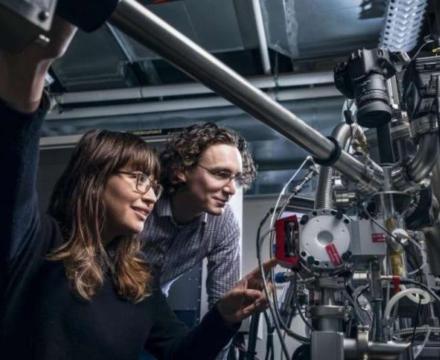 Two students adjusting and viewing a metal object, presumably a semiconductor, which consists of complex tubing and armatures with a black digital camera pointed down at it. One student appears to be adjusting the machine and pressing a button on a part of the machine painted red.