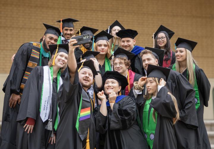 Graduates in caps and gowns take a celebratory selfie with a faculty member on commencement day