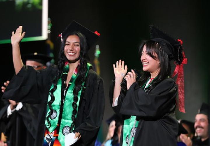 Two smiling graduates wave during the ceremony in cap and gown