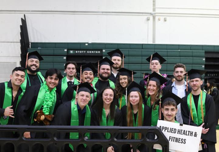 Group of Applied Health & Fitness graduates in caps and gowns, posing and smiling indoors