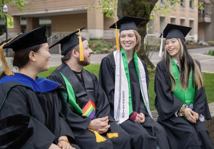 PSU graduates in caps and gowns sitting together and smiling on campus