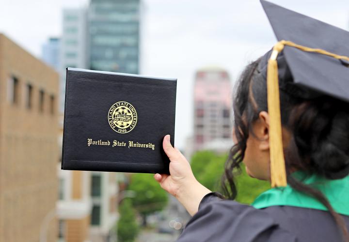 PSU graduate in cap and gown holding diploma cover overlooking downtown Portland