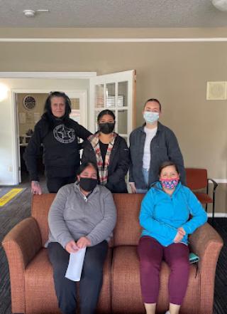 Five individuals staged around a red sofa smile for the camera, some with masks on.
