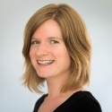 A headshot from the shoulders up of a smiling, white- and female presenting person with blonde, shoulder length hair.