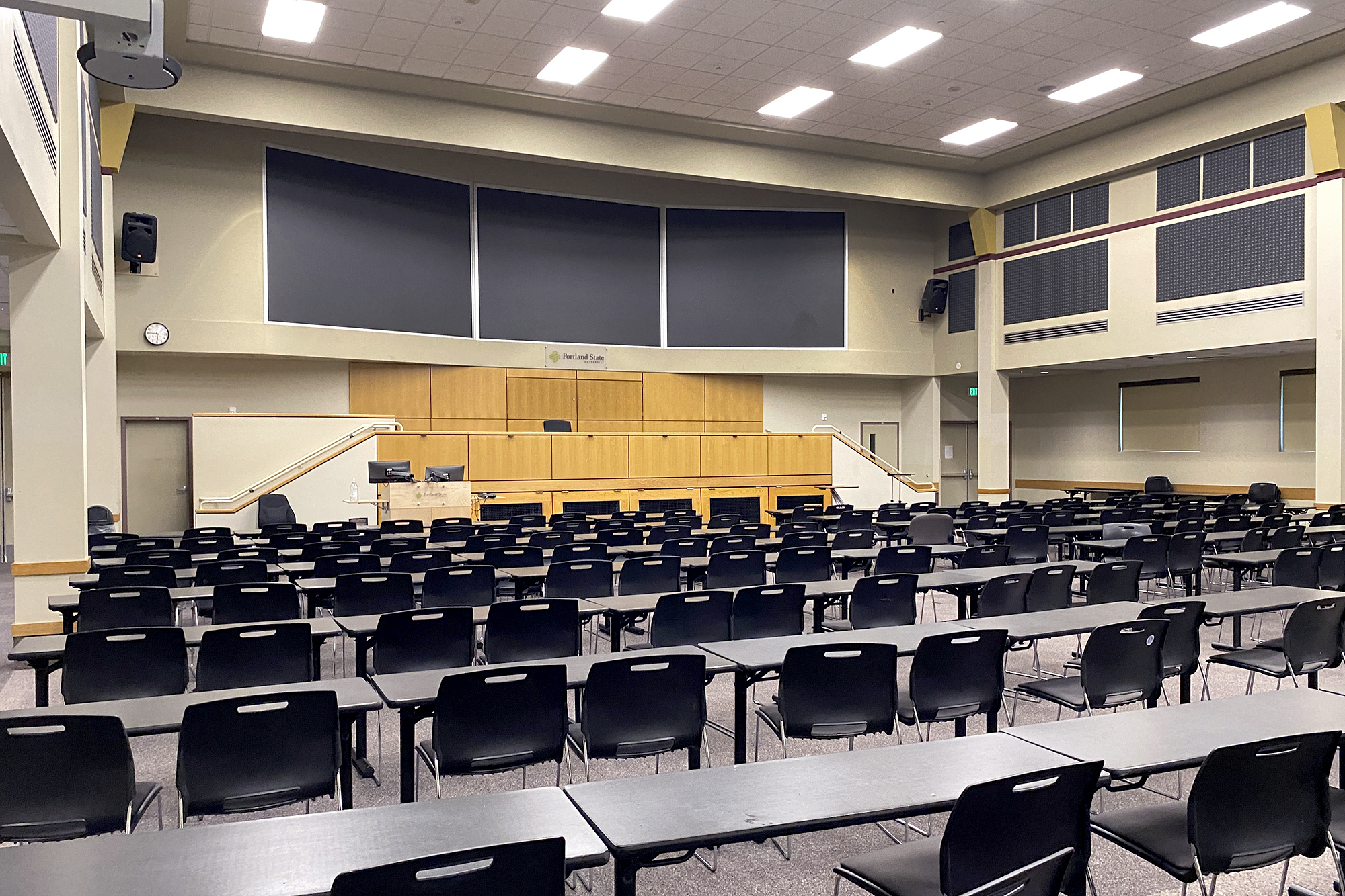 Hoffmann Hall consists of a main floor with a raised lectern platform, three large video screens above, computer-controlled audio and video equipment. Rows of chairs and class tables fill the floor.