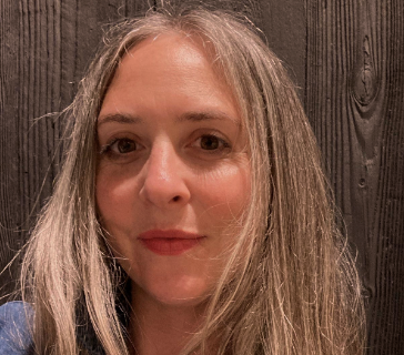 Photo of Rachel Toback. Rachel has blonde hair and is standing in front of a wood wall.