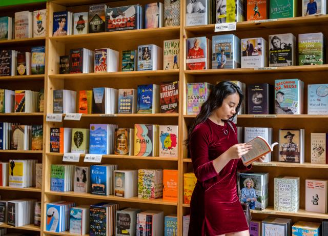 A person reading a book walks by a bookshelf