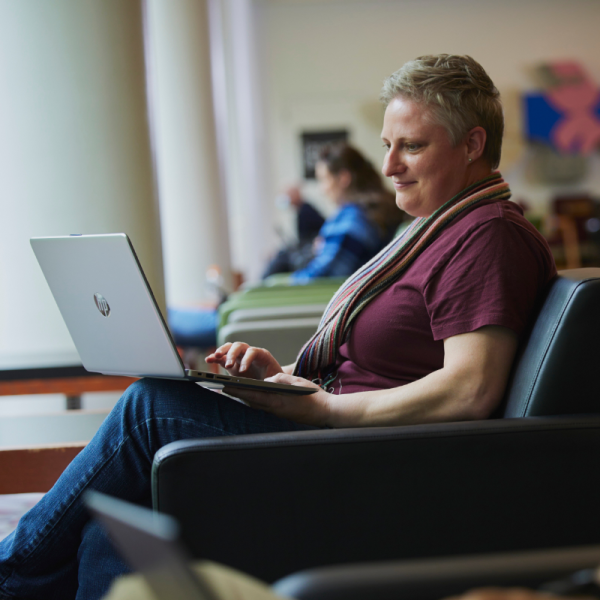 Photo of person sitting in comfortable chair with laptop on their knee. They are wearing a scarf and red t-shirt.