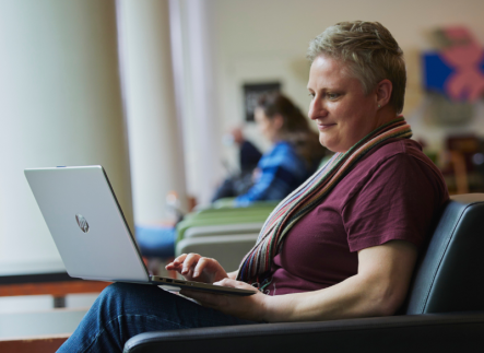 Photo of person sitting in comfortable chair with laptop on their knee. They are wearing a scarf and red t-shirt.