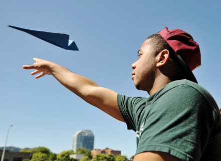 Photo of student wearing a red baseball cap and green t-shirt tossing a paper airplane into the air.