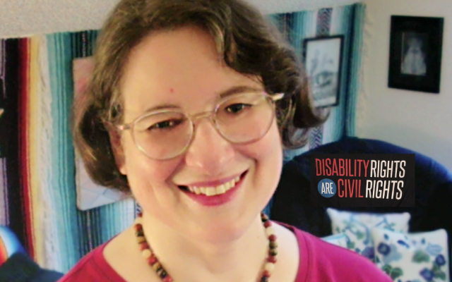 Darcy is smiling at the camera, wearing glasses, a red shirt, and beaded necklace. In the background "Disability Rights are Civil Rights" can be seen.