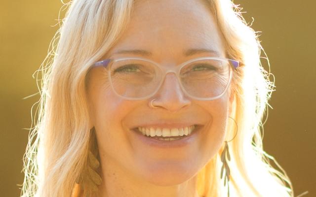 Photo of Abby - a blonde femme person smiling at the camera and wearing glasses.