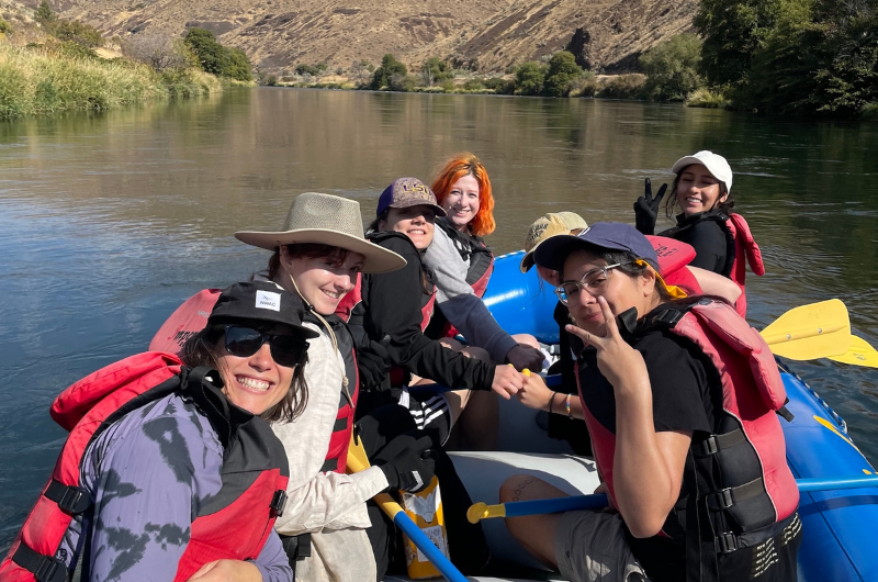 A group of Portland State students in life vests pose and smile while sitting in a raft on the Deschutes River.