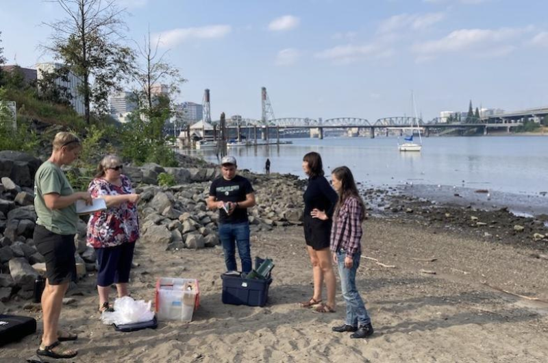 A group of researchers conduct fieldwork on a sandy riverbank, with the Willamette River, bridges, and the city skyline visible in the background.