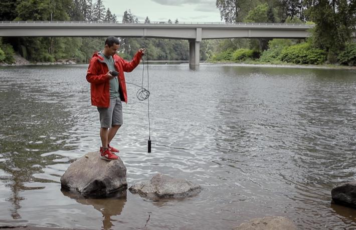 Student stands on a rock in a river, using equipment to test water quality.