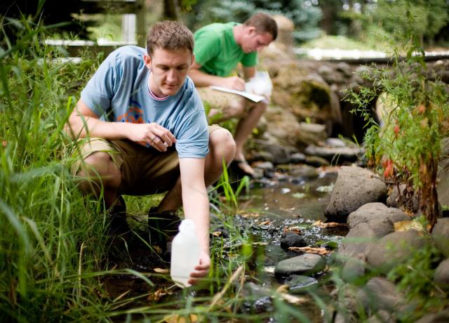Male student taking a water sample from a stream. Another male student is in the background making notes.