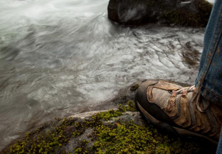 Feet standing on a moss-covered stone in a stream with three stones behind it.
