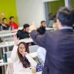 Students in a classroom, some with raised hands, and the back of an instructor calling on someone.