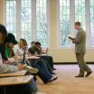 Instructor in front of a classroom standing and holding a book with front row of students taking notes