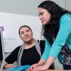 Two female students interacting in front of a computer, one looking up at the other and one looking at the monitor.