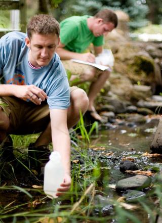 Male student taking a water sample from a stream. Another male student is in the background making notes.