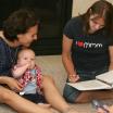 Two women sitting on the floor, one holding a baby while the other one writes in a notebook.