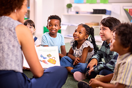 A diverse group of elementary school students sit on the floor watching their teacher read to them.