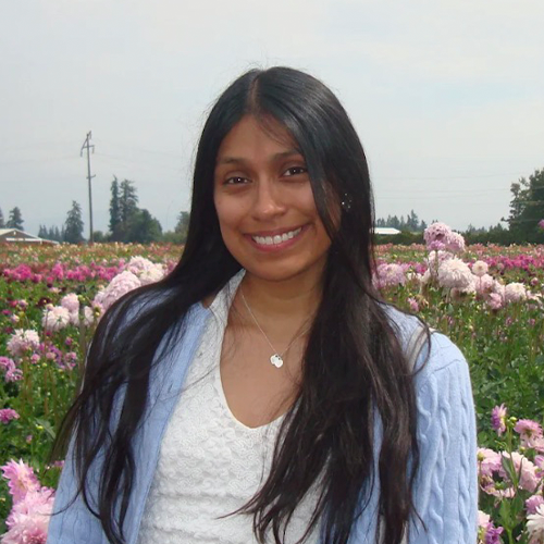 Stephani Jeronimo, academic advisor, is a young woman with brown skin and long black hair. She stands before a field of pink flowers.