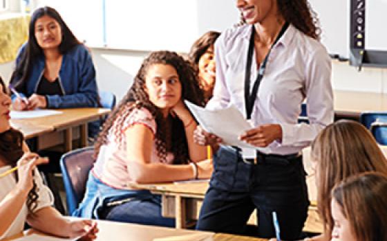 A teacher stands in the middle of a secondary school classroom talking with her students.