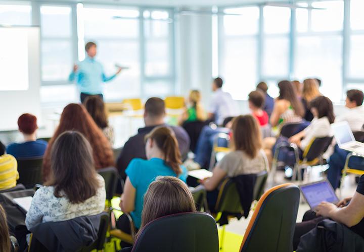 A blurry teacher across the room addresses a classroom full of students.