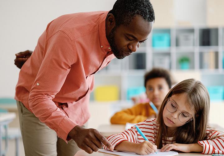 A Black male teacher in a salmon button-down and khaki slacks assists a student with her classwork.