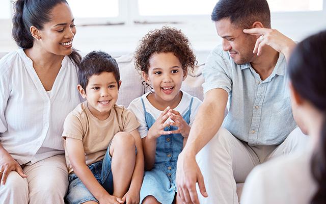 A mother, father, and two young children sit on a white couch together, smiling as they address a counselor.