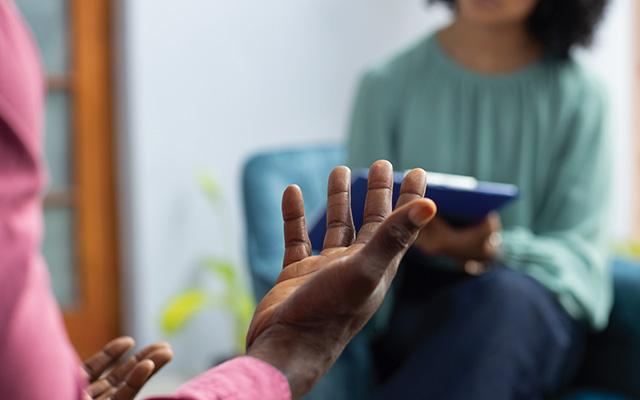 A counselor in a turquoise shirt holds a tablet and speaks with their patient, a man with dark skin and a pink shirt. Neither of their faces are visible.