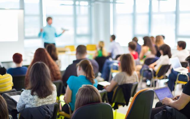 A blurry teacher across the room addresses a classroom full of students.