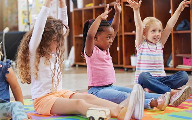 Several young children sit on their classroom floor with their arms up.