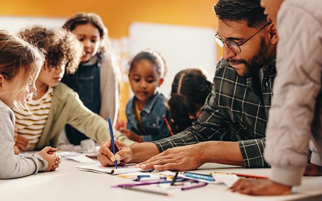 A teacher in a plaid shirt and glasses shows students how to complete an assignment.