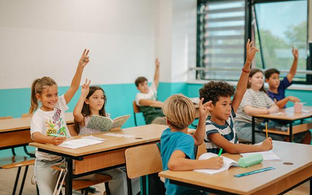 A white and teal classroom is full of students with their hands raised.