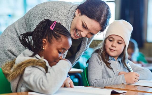 A Caucasian teacher with dark brown hair pulled back assists two students with their classwork.