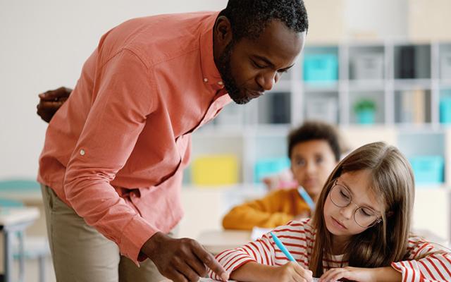 A Black male teacher in a salmon button-down and khaki slacks assists a student with her classwork.