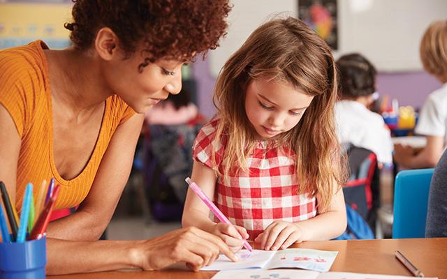 A teacher with curly, dark auburn hair helps a young girl in a red and white gingham top with schoolwork.