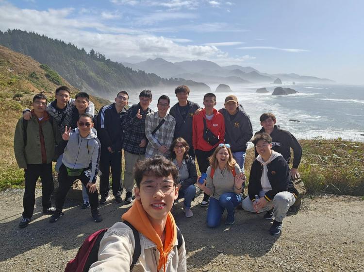 A large group of students posing for a selfie on a coastal cliffside, with a scenic view of the ocean and misty mountains in the background.
