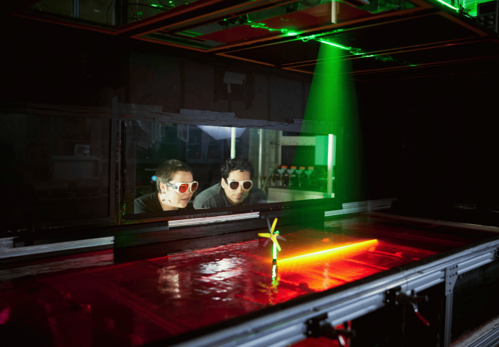 Two students wearing protective glasses watch a laser pointed at a miniature wind turbine over a tub of water.